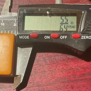 May include: A rectangular, amber-colored object is measured with a digital caliper. The digital display shows a measurement of 5.5 inches. The caliper is black with red buttons and a silver adjustment wheel. The object is placed on a dark wooden surface.