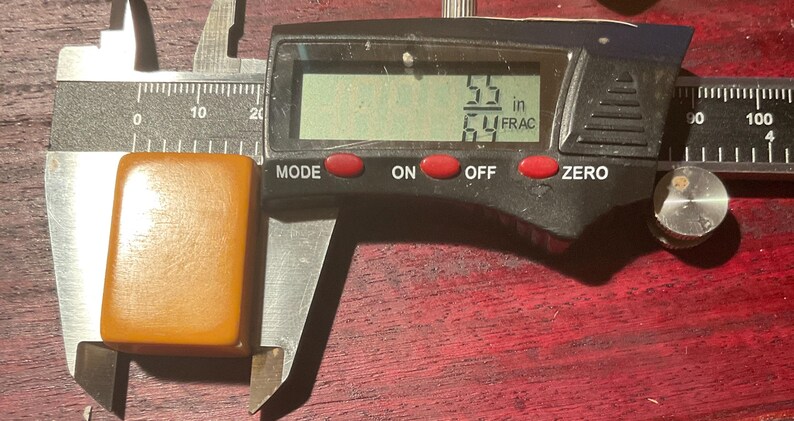 May include: A rectangular, amber-colored object is measured with a digital caliper. The digital display shows a measurement of 5.5 inches. The caliper is black with red buttons and a silver adjustment wheel. The object is placed on a dark wooden surface.