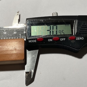 May include: A rectangular, light brown wooden block is positioned within the jaws of a black digital caliper. The digital display on the measuring tool clearly shows a measurement of 31.95 mm, indicating the length of the wooden object.