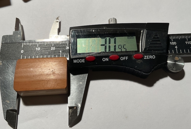 May include: A rectangular, light brown wooden block is positioned within the jaws of a black digital caliper. The digital display on the measuring tool clearly shows a measurement of 31.95 mm, indicating the length of the wooden object.