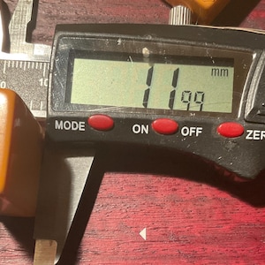 May include: A digital caliper measuring a rectangular, amber-colored object. The digital display shows 11.99 mm. The caliper is silver and black, with red buttons labeled "MODE", "ON", and "OFF".