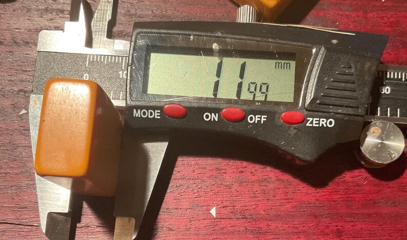 May include: A digital caliper measuring a rectangular, amber-colored object. The digital display shows 11.99 mm. The caliper is silver and black, with red buttons labeled "MODE", "ON", and "OFF".