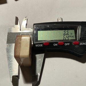 May include: A rectangular, two-toned beige and brown tile being measured by a black digital caliper. The digital display clearly shows a measurement of 33/64 inches. The tile has a smooth, solid appearance with a distinct color-blocked design.