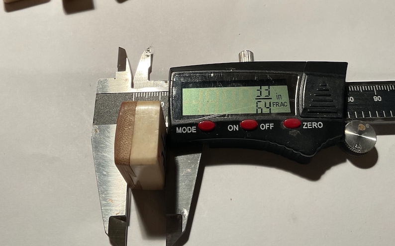 May include: A rectangular, two-toned beige and brown tile being measured by a black digital caliper. The digital display clearly shows a measurement of 33/64 inches. The tile has a smooth, solid appearance with a distinct color-blocked design.
