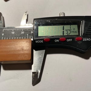 May include: A rectangular, light brown wooden block is positioned between the jaws of a black digital caliper. The digital display clearly shows a measurement of 1 1/32 inches, indicating the precise width of the wooden object being measured.
