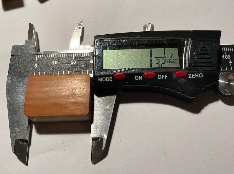 May include: A rectangular, light brown wooden block is positioned between the jaws of a black digital caliper. The digital display clearly shows a measurement of 1 1/32 inches, indicating the precise width of the wooden object being measured.