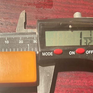 May include: A rectangular, amber-colored object is measured with a digital caliper. The caliper displays a measurement of 1.3 inches. The object is positioned on a dark, reddish-brown surface.