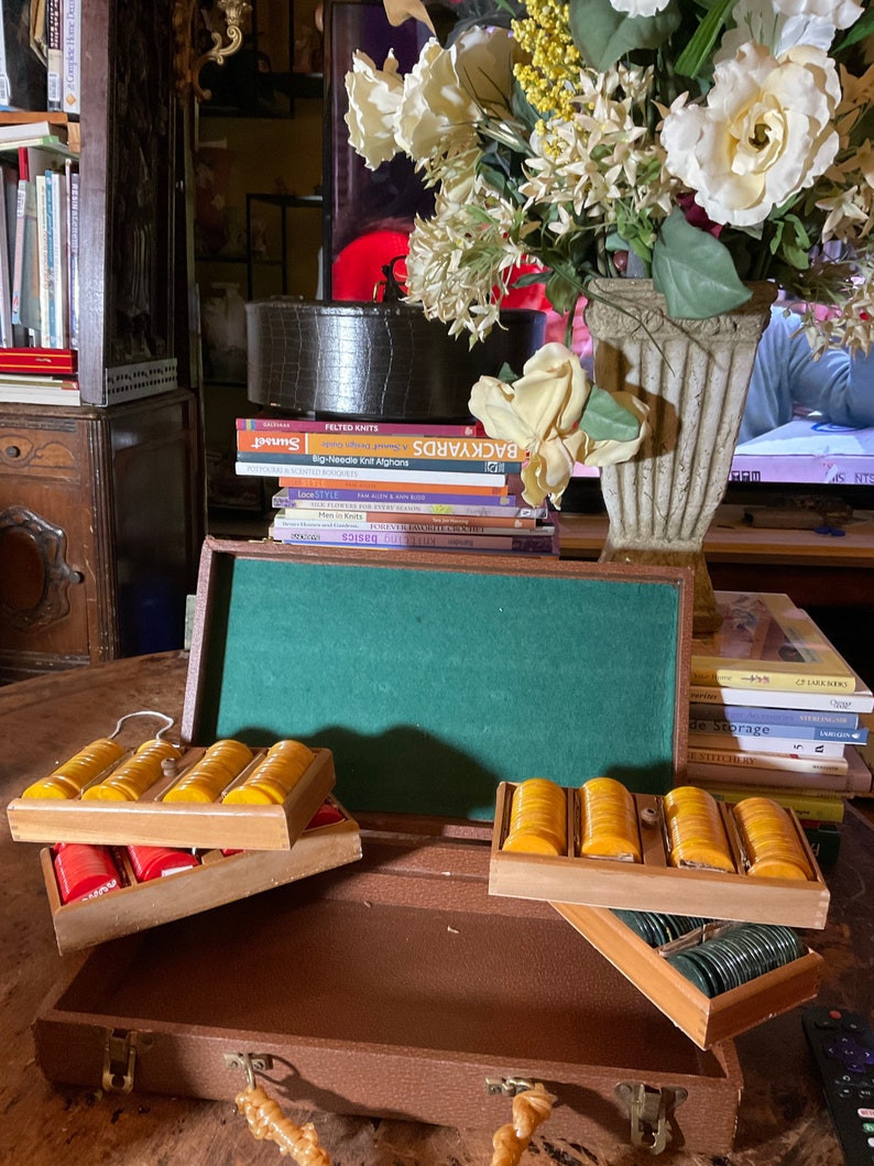 Pu&ograve; includere: Una valigetta vintage in similpelle marrone con interno in feltro verde, contenente diversi vassoi di fiches gialle, rosse e verdi. La valigetta &egrave; aperta, mostrando le fiches in vassoi di legno. Sullo sfondo &egrave; visibile una composizione floreale decorativa.