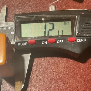 Puede incluir: Un calibrador digital mide un objeto rectangular de color &aacute;mbar. La pantalla muestra 12,11 mm. El calibrador es negro con botones rojos y una rueda de ajuste plateada. El objeto est&aacute; sobre una superficie marr&oacute;n rojiza.