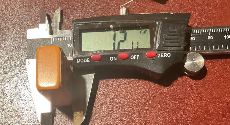 Puede incluir: Un calibrador digital mide un objeto rectangular de color &aacute;mbar. La pantalla muestra 12,11 mm. El calibrador es negro con botones rojos y una rueda de ajuste plateada. El objeto est&aacute; sobre una superficie marr&oacute;n rojiza.