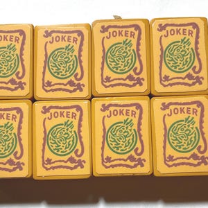 May include: Eight vintage Mahjong joker tiles. Each tile is rectangular with a yellow background, the word "JOKER" in brown, and a green and brown design. The tiles are arranged in two rows of four.