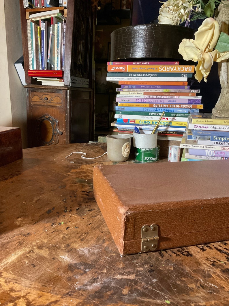 Pu&ograve; includere: Una custodia in pelle marrone con chiusura in ottone &egrave; appoggiata su un tavolo di legno. &Egrave; visibile una pila di libri, tra cui titoli come "Felted Knits" e "Backyards". Sul tavolo ci sono anche una piccola tazza di ceramica e un portapenne.