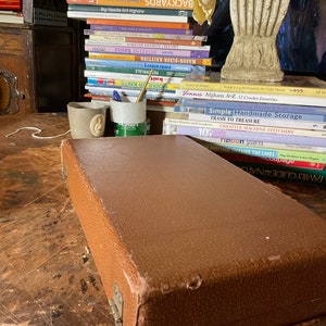 Pu&ograve; includere: Una custodia rettangolare marrone con superficie strutturata, appoggiata su un tavolo di legno. Sullo sfondo si vede una pila di libri sulla maglia e l'artigianato, insieme a un vaso decorativo e due piccole tazze di ceramica.