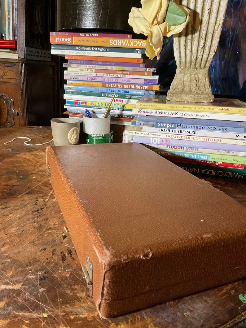 Pu&ograve; includere: Una custodia rettangolare marrone con superficie strutturata, appoggiata su un tavolo di legno. Sullo sfondo si vede una pila di libri sulla maglia e l'artigianato, insieme a un vaso decorativo e due piccole tazze di ceramica.