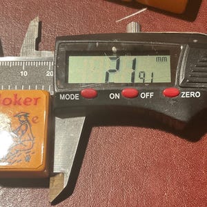 Puede incluir: Una ficha de juego rectangular, naranja, con la palabra "Joker" en rojo y una ilustraci&oacute;n en blanco y negro de una figura. La ficha se mide con un calibrador digital, mostrando un ancho de 19,1 mm y una longitud de 21 mm.