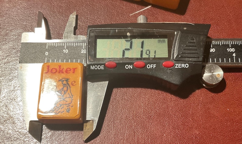 Puede incluir: Una ficha de juego rectangular, naranja, con la palabra "Joker" en rojo y una ilustraci&oacute;n en blanco y negro de una figura. La ficha se mide con un calibrador digital, mostrando un ancho de 19,1 mm y una longitud de 21 mm.