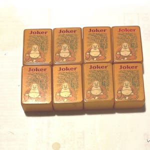 Puede incluir: Ocho fichas de juego rectangulares con una ilustración de Buda y la palabra "Joker" en rojo. Las fichas son de color marrón claro con un fondo de bambú.
