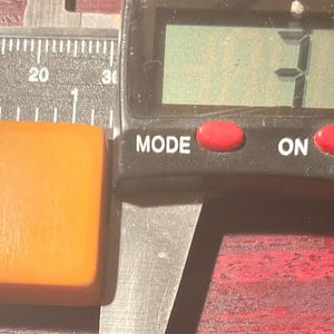 May include: An orange rectangular object is measured with a digital caliper. The object is approximately 31 mm long and 10.3 mm wide. The caliper has a digital display with red buttons for mode, on, off, and zero.