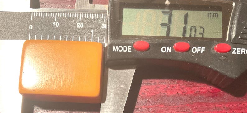 May include: An orange rectangular object is measured with a digital caliper. The object is approximately 31 mm long and 10.3 mm wide. The caliper has a digital display with red buttons for mode, on, off, and zero.