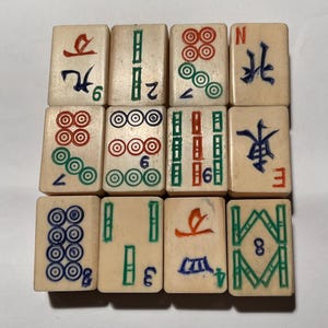 May include: A set of twelve off-white rectangular mahjong tiles arranged in a grid. Each tile features engraved symbols, characters, and numbers painted in red, green, blue, and black. The tiles display various suits including dots, bamboo, and winds.