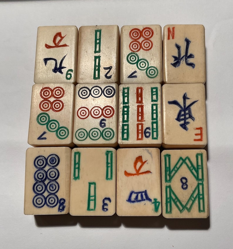 May include: A set of twelve off-white rectangular mahjong tiles arranged in a grid. Each tile features engraved symbols, characters, and numbers painted in red, green, blue, and black. The tiles display various suits including dots, bamboo, and winds.