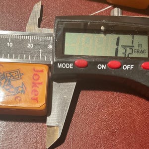 Puede incluir: Una ficha de Joker naranja de un juego, que mide 2,8 cm. La ficha tiene un gr&aacute;fico de una figura y la palabra "Joker" en rojo. Un calibrador digital muestra la medida.