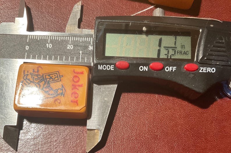 Puede incluir: Una ficha de Joker naranja de un juego, que mide 2,8 cm. La ficha tiene un gr&aacute;fico de una figura y la palabra "Joker" en rojo. Un calibrador digital muestra la medida.