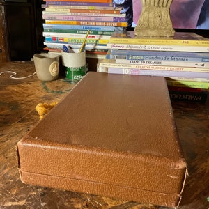 Pu&ograve; includere: Una scatola rettangolare marrone con una superficie strutturata &egrave; appoggiata su un tavolo di legno. La scatola &egrave; chiusa e sembra essere fatta di un materiale simile alla pelle. Sullo sfondo &egrave; visibile una pila di libri, che suggerisce un tema artigianale o hobbistico.
