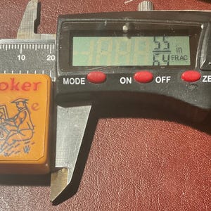 Puede incluir: Una ficha de juego rectangular vintage con la palabra "Joker" en rojo. La ficha presenta una ilustraci&oacute;n de una persona pintando. La ficha mide 2,18 cm de largo. Se utiliza un calibrador digital para medir la ficha.