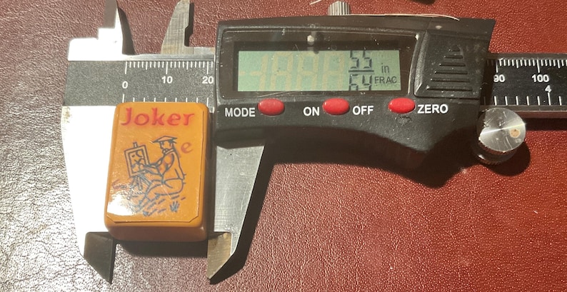 Puede incluir: Una ficha de juego rectangular vintage con la palabra "Joker" en rojo. La ficha presenta una ilustraci&oacute;n de una persona pintando. La ficha mide 2,18 cm de largo. Se utiliza un calibrador digital para medir la ficha.
