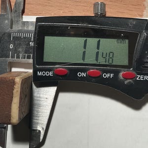 以下が含まれることがあります： デジタルノギスで小さな長方形の物体を測定しています。物体の幅は約1.14cmです。ノギスのデジタル表示はミリメートルで測定値を示しています。物体は茶色のテクスチャ加工された表面を持っています。