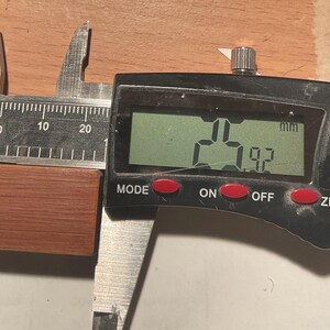 以下が含まれることがあります： デジタルノギスで、赤茶色の小さな長方形の木片を測定しています。デジタルディスプレイには、2.5 cm x 1.9 cmの測定値が表示されます。ノギスは黒色で、赤いボタンが付いています。