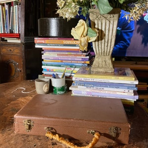 Pu&ograve; includere: Una valigia marrone con manico intrecciato e chiusure in ottone &egrave; appoggiata su un tavolo di legno. Una pila di libri sulla maglieria e l'artigianato &egrave; dietro la valigia. Un vaso di fiori e due piccole tazze sono anche sul tavolo.