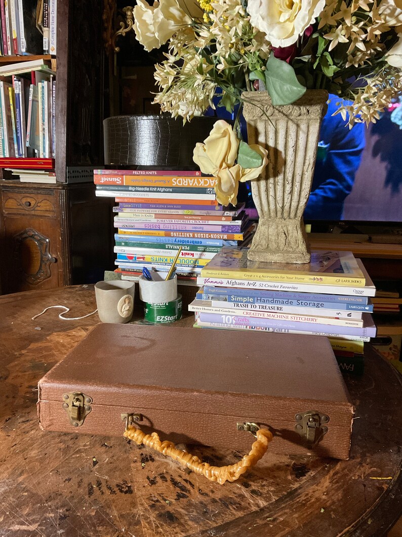 Pu&ograve; includere: Una valigia marrone con manico intrecciato e chiusure in ottone &egrave; appoggiata su un tavolo di legno. Una pila di libri sulla maglieria e l'artigianato &egrave; dietro la valigia. Un vaso di fiori e due piccole tazze sono anche sul tavolo.