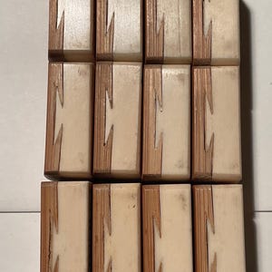 May include: A collection of twelve rectangular wooden blocks arranged in a grid of three rows and four columns. Each block features a two-tone design with a light cream-colored section joined to a textured brown wood section by a zigzag joint.