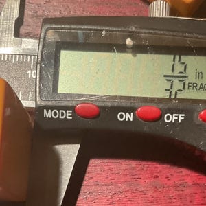 May include: An orange rectangular object is measured with a digital caliper. The digital display shows a measurement of 15/37 inches. The caliper is black and silver, with red buttons labeled "MODE", "ON", "OFF", and "ZERO".