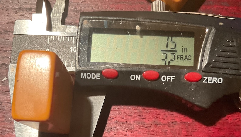 May include: An orange rectangular object is measured with a digital caliper. The digital display shows a measurement of 15/37 inches. The caliper is black and silver, with red buttons labeled "MODE", "ON", "OFF", and "ZERO".