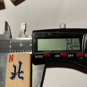 May include: A rectangular off-white mahjong tile featuring a blue engraved Chinese character and a red letter N at the top. The tile is positioned within a black digital caliper showing a measurement of 53/64 inches on the LCD screen display.