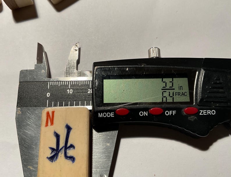 May include: A rectangular off-white mahjong tile featuring a blue engraved Chinese character and a red letter N at the top. The tile is positioned within a black digital caliper showing a measurement of 53/64 inches on the LCD screen display.