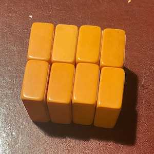 Puede incluir: Ocho baldosas rectangulares de color amarillo anaranjado dispuestas en dos filas de cuatro. Las baldosas tienen bordes redondeados y un acabado liso y brillante. El fondo es una superficie marr&oacute;n rojizo oscuro.