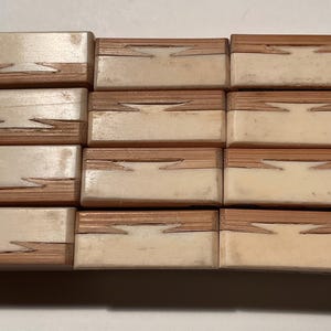 May include: A collection of twelve rectangular blocks arranged in a grid, each featuring a two-tone design with a light cream-colored section and a natural wood-grain section joined by a distinct dovetail-style interlocking joint pattern.