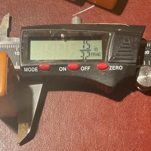 Puede incluir: Un calibrador digital que mide un objeto rectangular de color caramelo. La pantalla digital muestra 3,8 cm. El calibrador tiene botones rojos etiquetados "MODE", "ON", "OFF" y "ZERO". El objeto est&aacute; sobre una superficie roja.