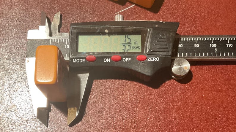 Puede incluir: Un calibrador digital que mide un objeto rectangular de color caramelo. La pantalla digital muestra 3,8 cm. El calibrador tiene botones rojos etiquetados "MODE", "ON", "OFF" y "ZERO". El objeto est&aacute; sobre una superficie roja.