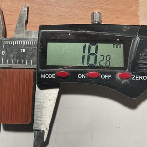 以下が含まれることがあります： 赤茶色の長方形の物体で、幅約1.8cm。デジタルノギスで測定されています。ノギスは18.28mmの測定値を表示します。物体はテクスチャ加工された、溝のある表面を持っています。