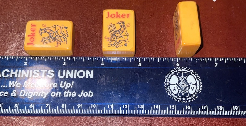 Puede incluir: Tres fichas de Mahjong amarillas vintage con texto "Joker" e ilustraciones. Una muestra un perro, otra un artista y la tercera est&aacute; en blanco. Una regla debajo mide en cent&iacute;metros.
