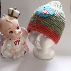 Puede incluir: Un gorro de bebé de crochet verde y naranja con una aplicación de fieltro azul en forma de pájaro. El pájaro tiene un corazón de fieltro blanco con lunares rosas y blancos en el pecho.