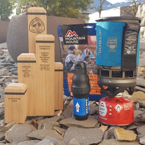 Könnte beinhalten: Ein blaues Jetboil Flash Kochsystem mit einer roten Kartusche, einer schwarzen und blauen Wasserflasche und einem Mountain House gefriergetrockneten Mahlzeitenbeutel. Die Gegenstände befinden sich auf einem Bett aus grauen Steinen.