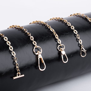 May include: Five gold-colored chain straps with different clasps and hooks. The chains are laid out on a black leather surface.