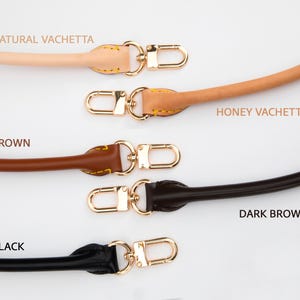 May include: Five leather bag straps in natural vachetta, honey vachetta, brown, dark brown, and black. Each strap features a gold-coloured clasp. The text "NATURAL VACHETTA", "HONEY VACHETTA", "BROWN", "DARK BROWN", and "BLACK" are printed above or below each strap.