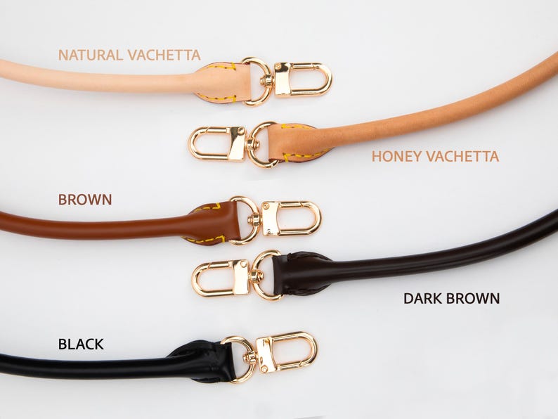 May include: Five leather bag straps in natural vachetta, honey vachetta, brown, dark brown, and black. Each strap features a gold-coloured clasp. The text "NATURAL VACHETTA", "HONEY VACHETTA", "BROWN", "DARK BROWN", and "BLACK" are printed above or below each strap.